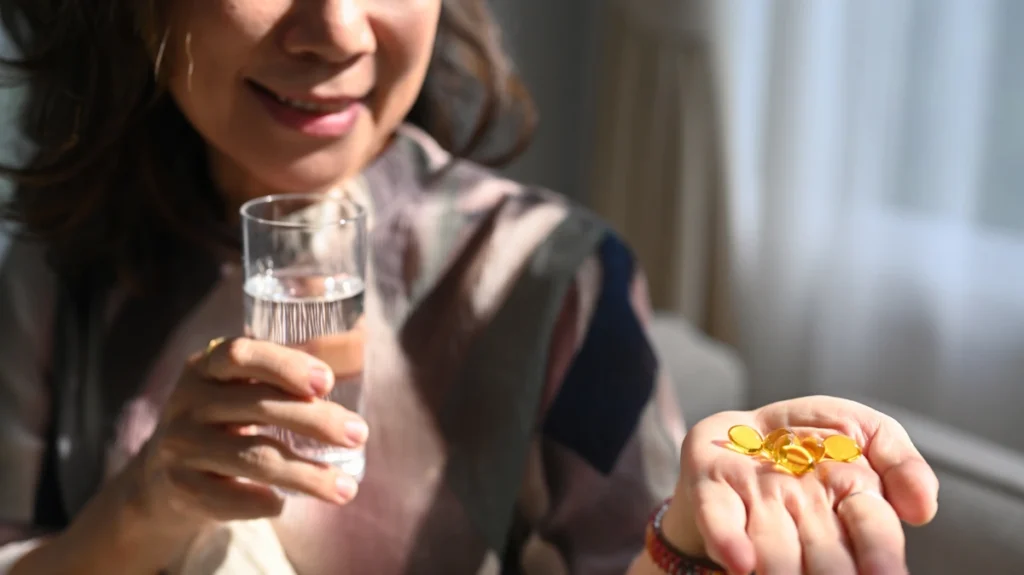 Une femme qui tient un verre d'eau dans une main et des gélules jaune dans l'autre